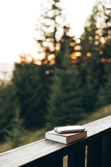 Smartphone and paper Bible on wooden railing, forest background, morning light.