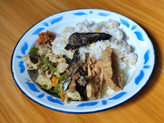 A portion of typical Indonesian mixed rice served on a classic blue and white patterned enamel plate, consisting of corn rice, a piece of dried fish, fried tempeh, stir-fry and chili sauce. Delicious 