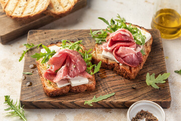 Grilled toasts with ham, cheese and arugula on wooden cutting board on light textured background. Delicious breakfast or snack