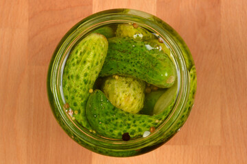 A top-down view of a glass jar filled with fresh pickling cucumbers and spices, resting on a wooden...
