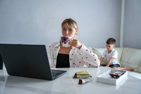Mother working on laptop at home while son plays on sofa in the background - Powered by Adobe
