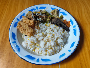 A portion of typical Indonesian mixed rice served on a classic blue and white patterned enamel plate, consisting of corn rice, a piece of dried fish, fried tempeh, stir-fry and chili sauce. Delicious 