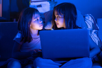 Two Asian girls sit on the sofa watching a laptop at night. Parental control, screen addiction, and...