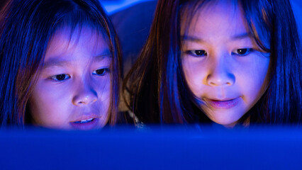Two Asian girls sit on the sofa watching a laptop at night. Parental control, screen addiction, and...