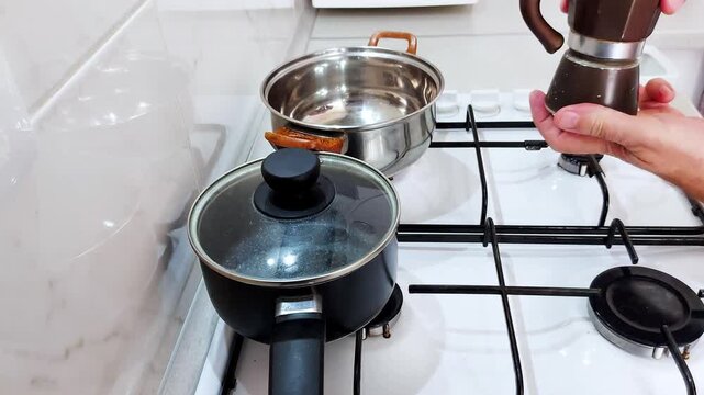 Hands adjust a small coffee maker on a clean gas stove beside polished pots and pans-capturing a quiet kitchen moment of preparation, warmth, and everyday rhythm