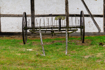 Antique horse-drawn hay rake with iron wheels, standing on grass in front of an old half-timbered...