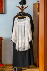 Traditional Catholic priest's attire hanging on a coat rack – black cassock, white surplice, and biretta, displayed in a classic interior