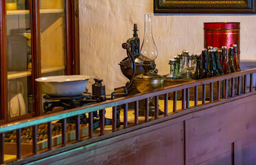 Interior of an old tavern with vintage kitchenware, a kerosene lamp, and a collection of glass bottles arranged on a wooden sideboard. Rustic atmosphere with historical charm.