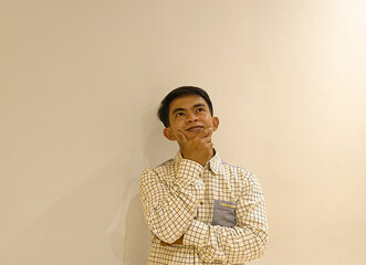 Fototapeta premium A young Asian man in a checkered shirt standing against a plain beige wall with a thoughtful expression