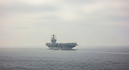 Massive grey aircraft carrier underway on a vast, calm ocean under a hazy, overcast sky, showcasing naval power and maritime operations.