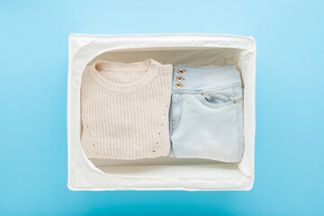 Knitted sweater and jeans in white textile storage case on light blue table background. Pastel color. Female clothes. Closeup. Top down view.
