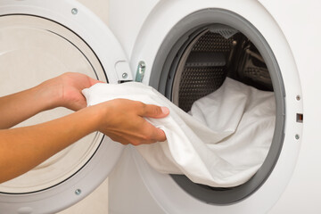 Young adult woman hands holding and unloading white sheet from opened washing machine. Clothes care. Closeup. Side view.