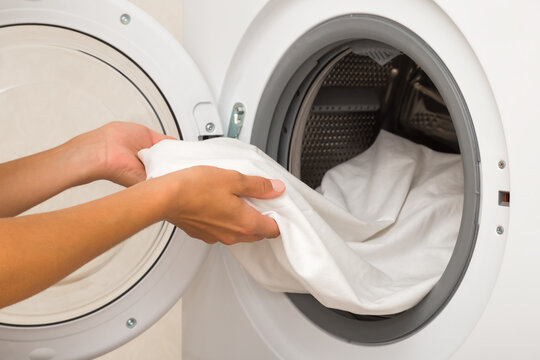 Young adult woman hands holding and unloading white sheet from opened washing machine. Clothes care. Closeup. Side view.