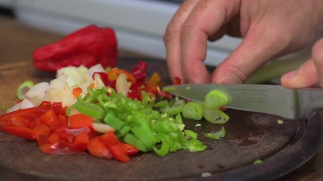Close up of hands cutting scallion, pepper, and onion on wooden board in kitchen