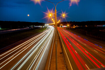 Obraz premium Traces of white and red stripes from the headlights of cars on the highway at night. Transport construction industry.