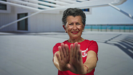 Senior woman with grey hair in red shirt gesturing stop at seaside promenade during daylight,...