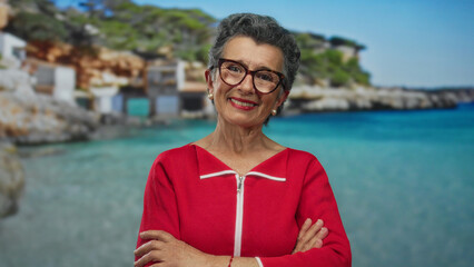 Senior woman with grey hair in a red sweater smiling with crossed arms on a sunny seaside promenade...