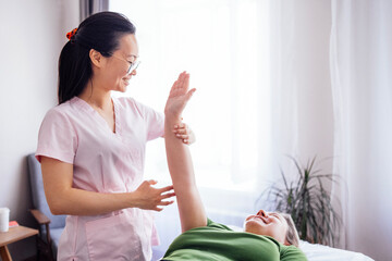 Female therapist in pink scrubs is providing physical therapy to a smiling woman in green shirt,...