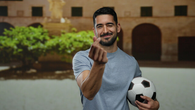 Young man making heart gesture with fingers while holding a soccer ball on a city street showing a playful and cheerful expression.