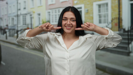 Fototapeta premium Young plus size woman smiles and points at her smile with fingers while standing confidently outdoors on a city street during daytime.