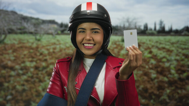 Young woman in helmet and red jacket with arm in sling holding credit card outdoors in green park setting.