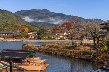 Naklejka premium Wooden boats rest on calm Katsura River in Arashiyama, Kyoto, Japan, as morning mist floats over colorful hills and traditional houses along quiet riverside
