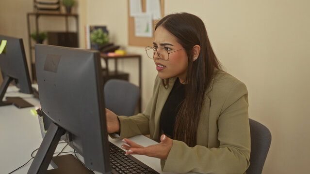 Woman in office wearing glasses and blazer appears frustrated while staring at computer screen in workplace setting, suggesting stress or confusion in a professional environment.
