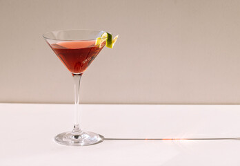 Close-up of a martini glass on a white table. An elegant goblet with a red alcoholic sparkling drink decorated with lime zest. Tasty berry cocktail.