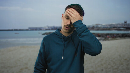 Young hispanic man in distress rubbing forehead at a sunny seaside beach with a scenic city...