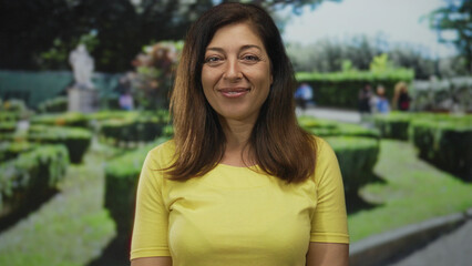 Middle age woman smiling beside manicured hedges in a green park under sunlight near stone statue; serenity calm joy peace.