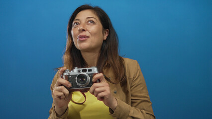 Middle aged hispanic woman holding vintage camera with bare hands in blue professional studio environment; curiosity.