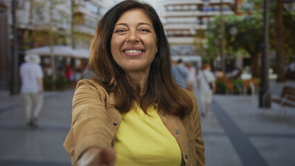 Woman in casual brown jacket with warm smile extends hand toward camera on bustling street;...
