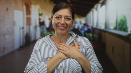 Middle aged hispanic woman in striped shirt smiling with hands on chest in sunlit building;...