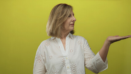 Middle age woman wearing a white blouse points finger to open palm against a yellow backdrop in...