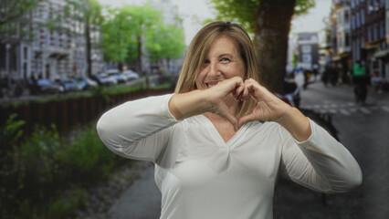 Middle aged caucasian woman smiling and forming heart with hands on urban street; love happiness...