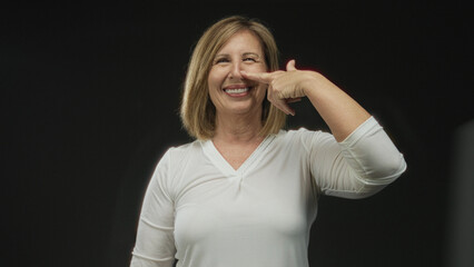 Woman smiling while pointing finger to nose in black studio wearing white long sleeve top;...