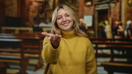 Woman smiling in warm sweater inside a church setting extending hand invitingly with wooden pews...