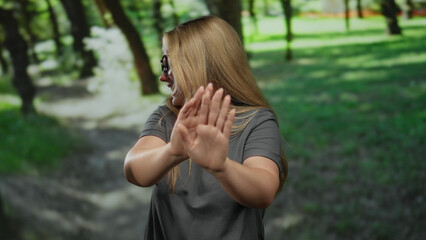 Woman gestures stop with hands outdoors in park surrounded by lush green trees, exuding youthful...