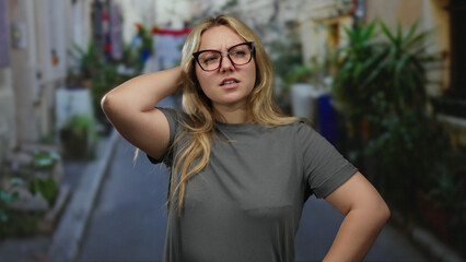 Woman standing on city street with glasses and blonde hair in casual clothing outdoors with thoughtful expressions on an urban backdrop.