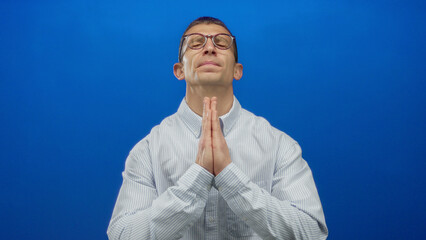 Caucasian man wearing glasses and striped shirt praying with hands clasped against blue isolated...