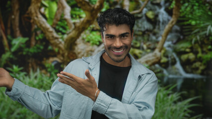 Young man in light jacket pointing hand in lush green park by waterfall, smiling broadly forward;...