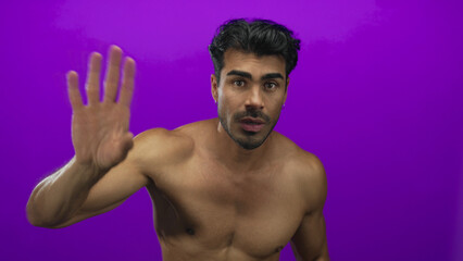 Fototapeta premium Young hispanic man with muscular bare chest points fingers downward in purple studio; invitation.