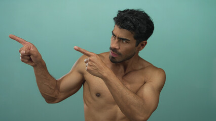 Shirtless young hispanic man pointing both index fingers left with furrowed brow in teal studio;...