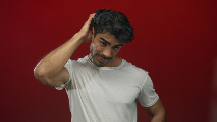 Man in white t-shirt scratching his head in a red studio setting while looking down with puzzled...