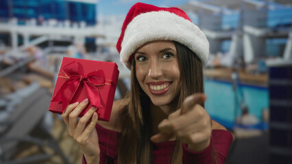 Young hispanic woman points finger to camera holding red gift box on cruise ship deck; holiday...