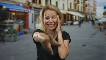 Woman with blonde hair in black shirt smiles broadly and points finger at camera on busy street;...