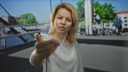 Woman in gray blouse standing on urban street beside canal railing points finger; assertion confidence.