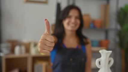 Artisan woman holds clay vase in one hand and shows thumb up with other hand in studio filled with...