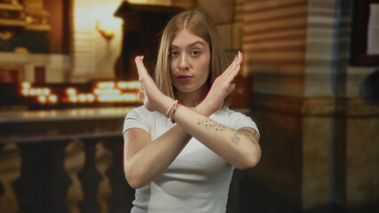 Young blonde woman with arms crossed in a candlelit gothic church hall stop gesture; denial rejection.