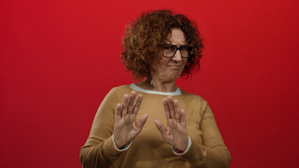 Hispanic woman expressing rejection with a gesture and frown, standing against a vivid red wall,...